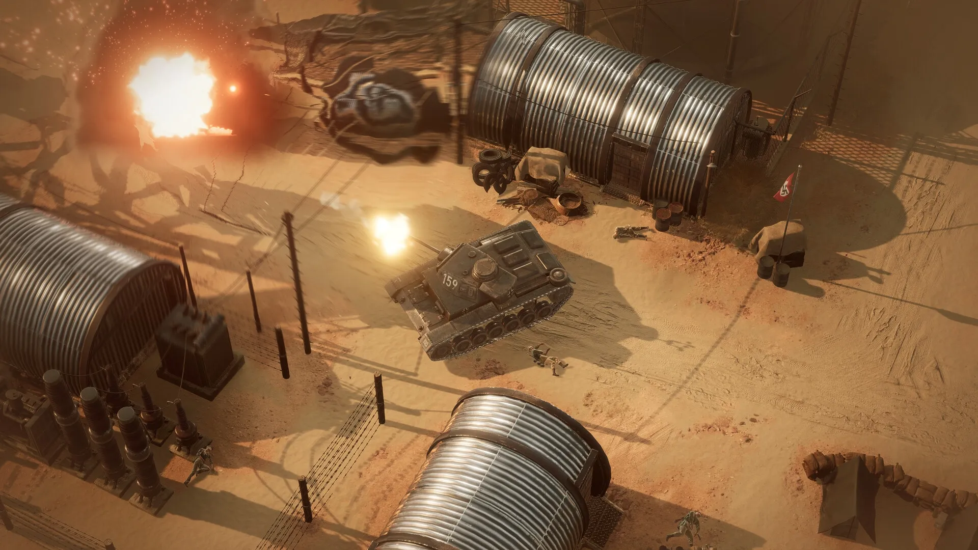 Commandos: Tank firing near explosion in desert military base with multiple metal huts and barbed wire.