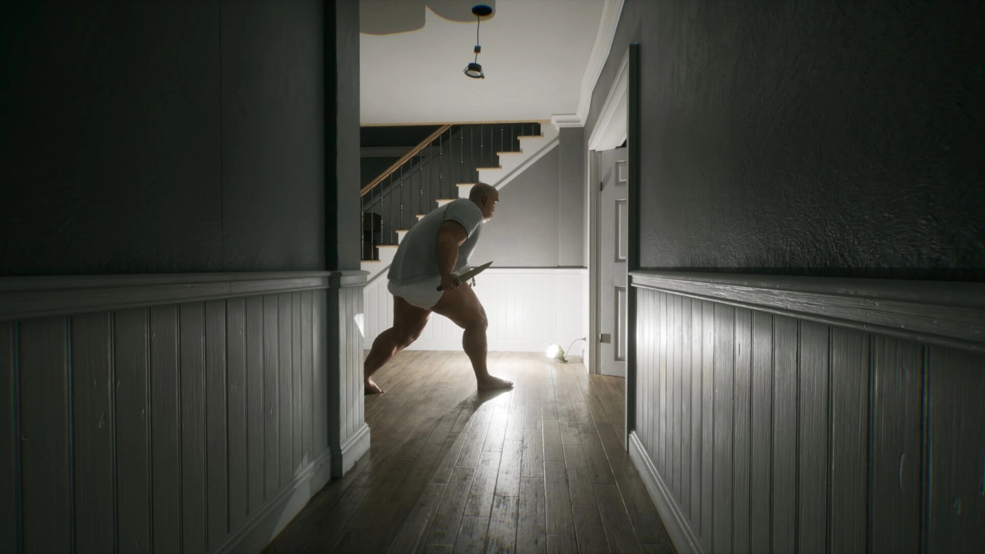 LAG KILLS: Barefoot man crouch-walks with a knife in a dim hallway near stairs and door light.