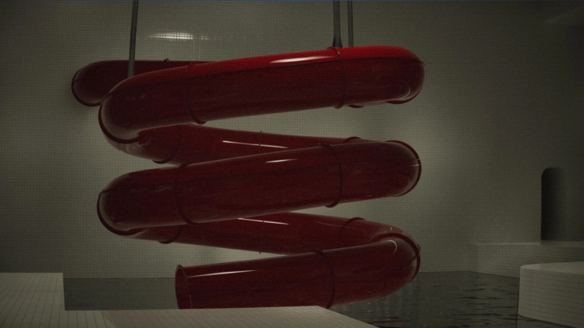 Late Shift: View of a red spiral tube slide inside a dim tiled pool room with still water below.