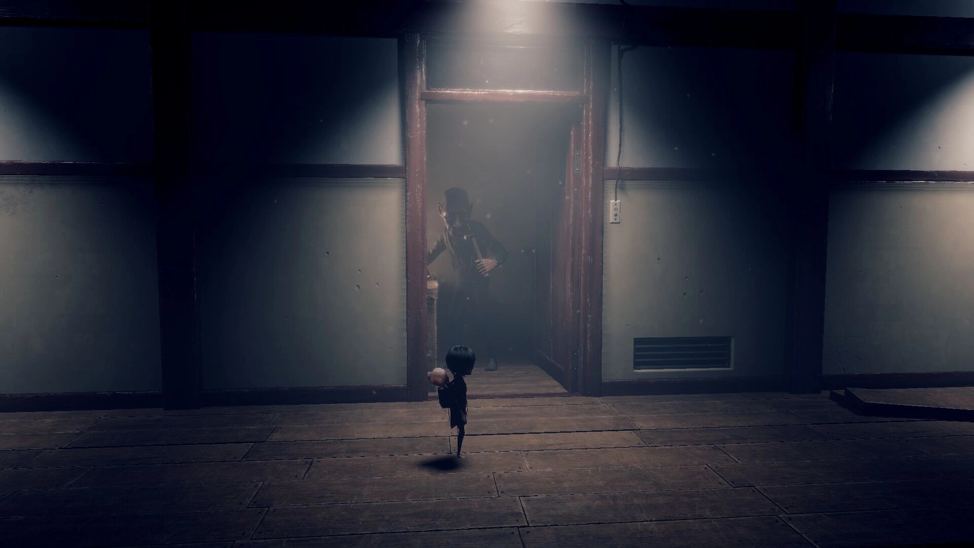 Lull: Girl carrying baby runs from shadowy figure through doorway in dim, dusty facility corridor.