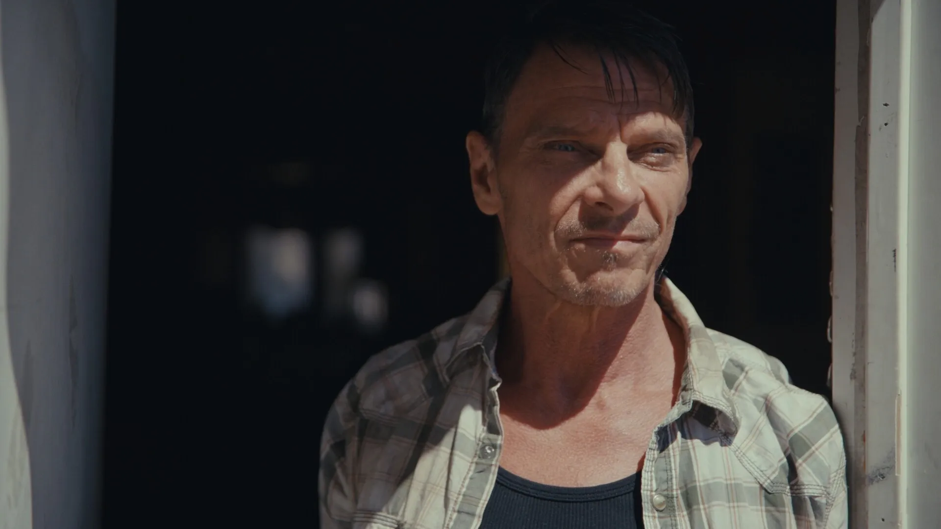 Nowhere to Run: Man standing in dimly lit doorway, expression serious, wearing plaid shirt and black tank top