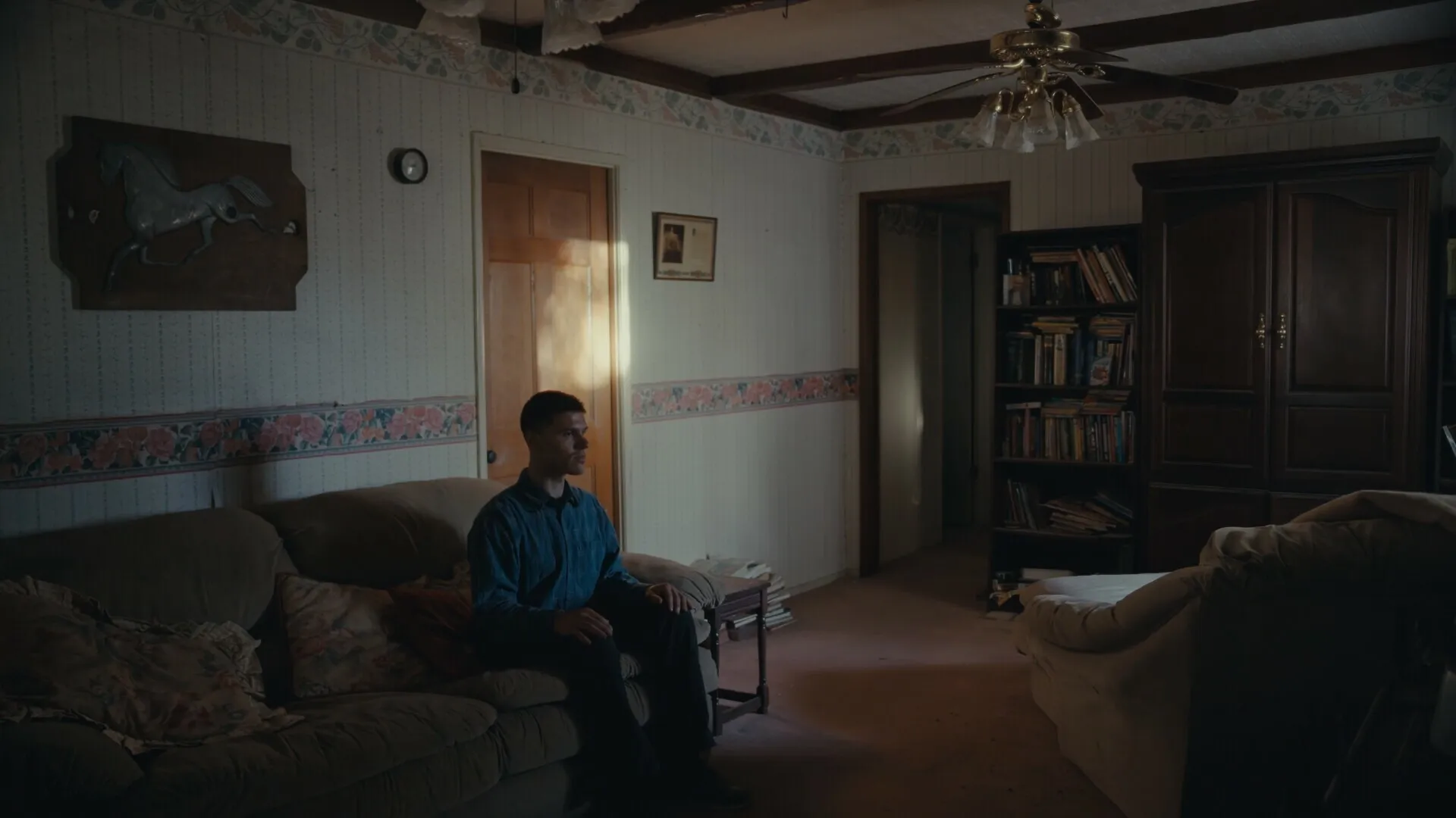Nowhere to Run: Man sits on a couch in a dimly lit living room with bookshelves and wooden decor