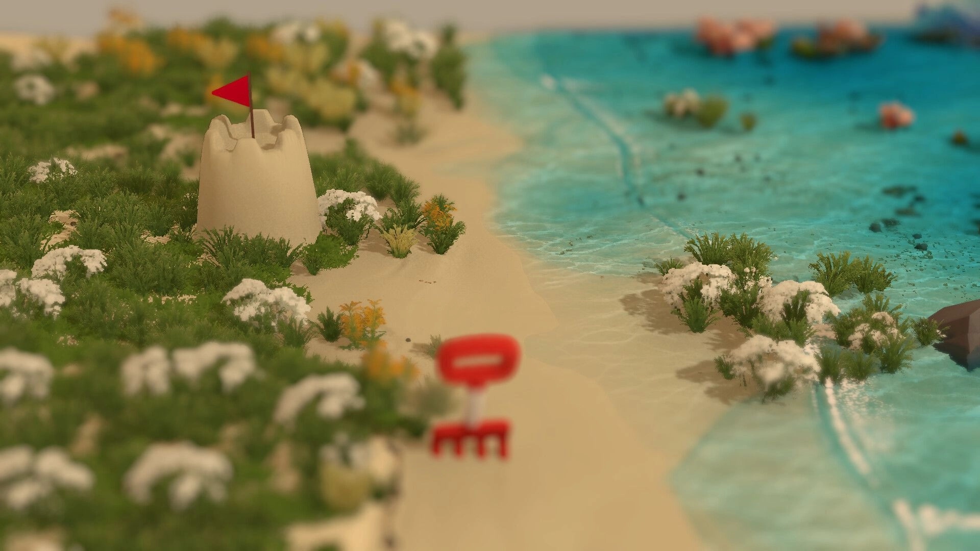 Sandcastle: Sand tower with red flag on grassy beach beside turquoise water, blurred rake icon UI.
