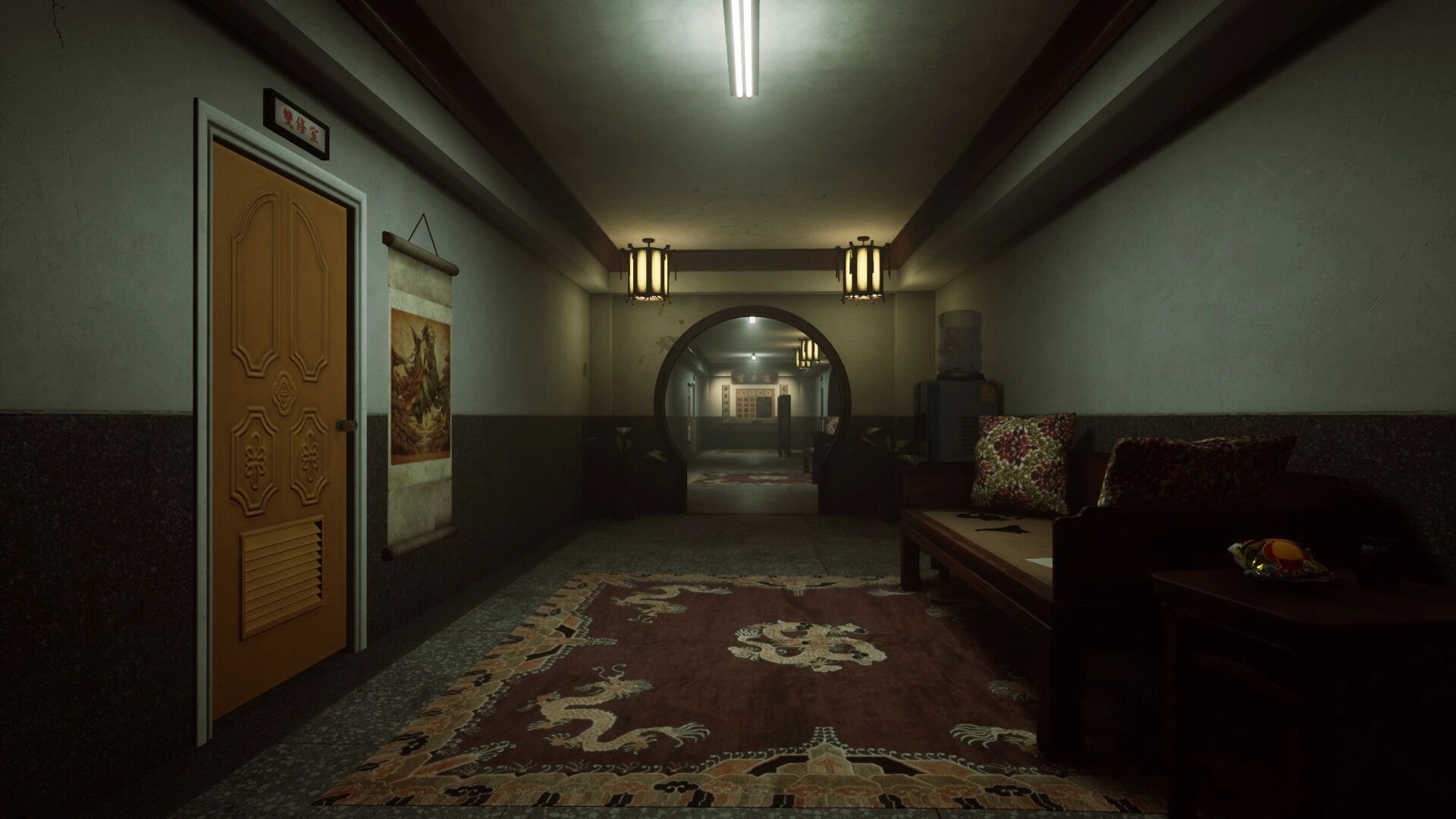 SUFFOCATE: Dimly lit hallway with ornate carpet, closed door, sofa, and wall scroll in apartment setting.